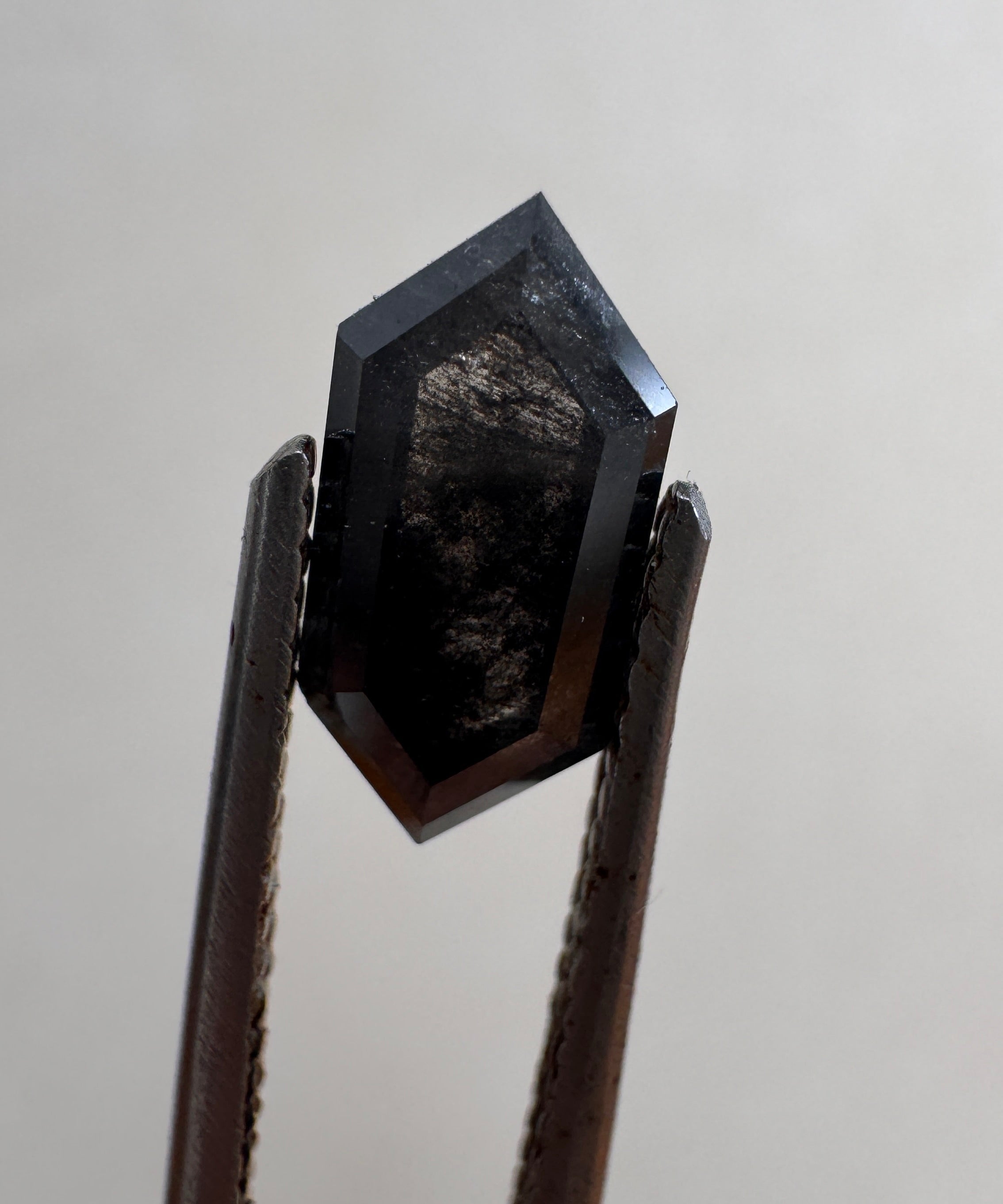 Close-up of a 1.36ct natural black elongated hexagon step cut diamond held in tweezers, showcasing its striking dark geometric facets. GIA certified and sourced for MOHSTEN bespoke custom engagement rings in Auckland, New Zealand.