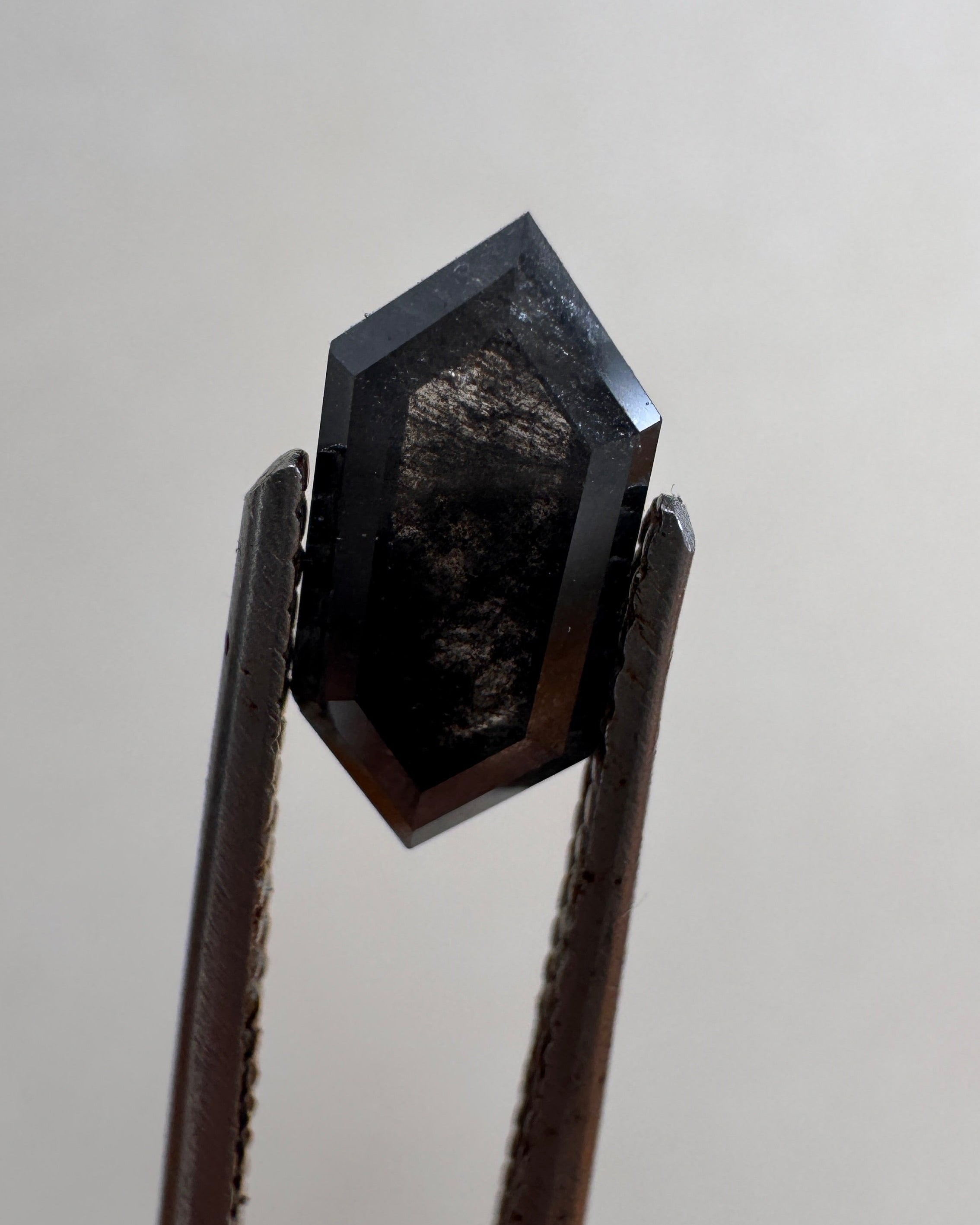 Close-up of a 1.36ct natural black elongated hexagon step cut diamond held in tweezers, showcasing its striking dark geometric facets. GIA certified and sourced for MOHSTEN bespoke custom engagement rings in Auckland, New Zealand.
