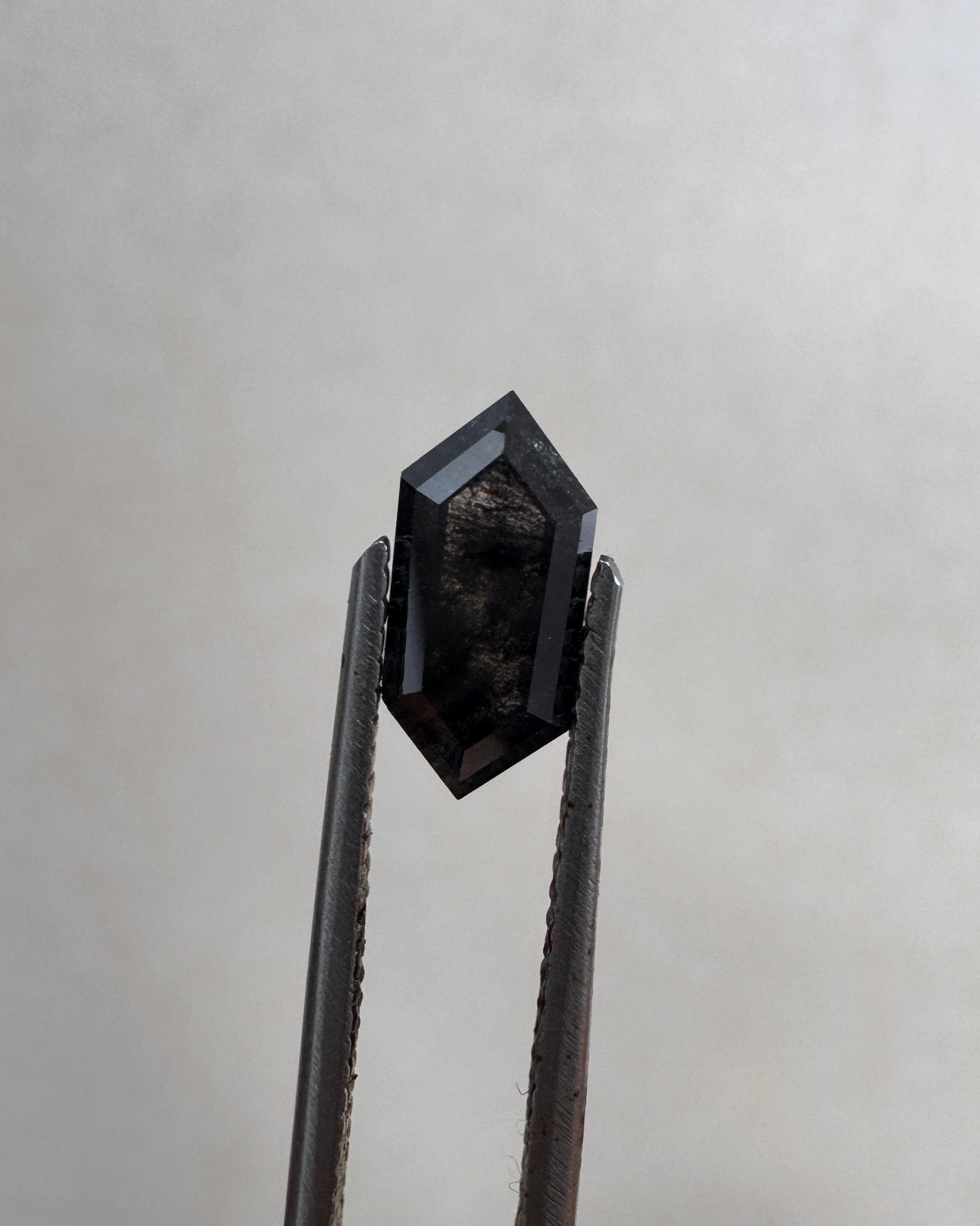 Close-up of a 1.36ct natural black elongated hexagon step cut diamond held in tweezers, showcasing its striking dark geometric facets. GIA certified and sourced for MOHSTEN bespoke custom engagement rings in Auckland, New Zealand.