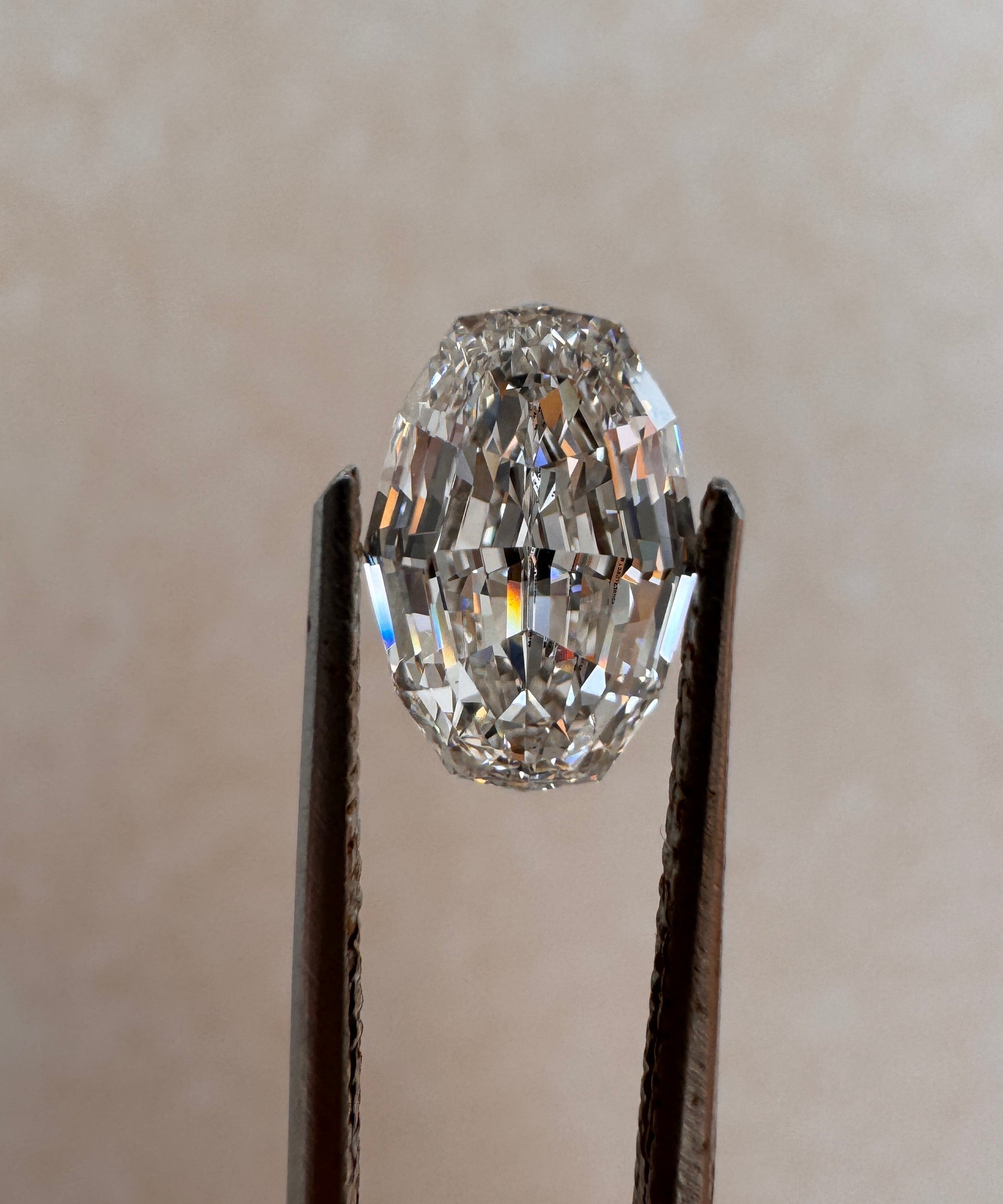 A loose 2.09ct oval step cut lab-grown diamond held upright by metal tweezers against a soft beige background. Sourced for bespoke custom engagement rings by MOHSTEN jewellery studio in Auckland, New Zealand.