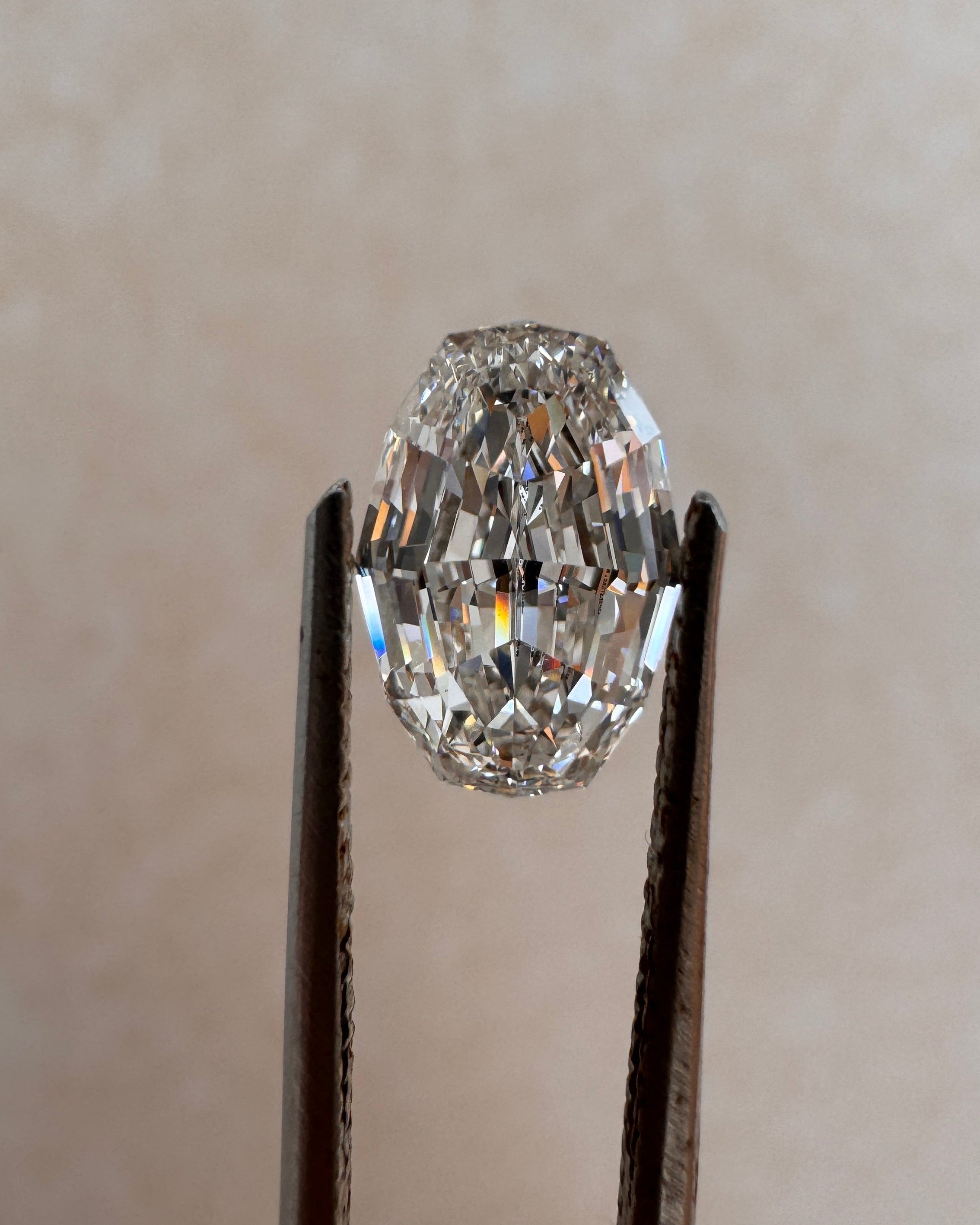 A loose 2.09ct oval step cut lab-grown diamond held upright by metal tweezers against a soft beige background. Sourced for bespoke custom engagement rings by MOHSTEN jewellery studio in Auckland, New Zealand.