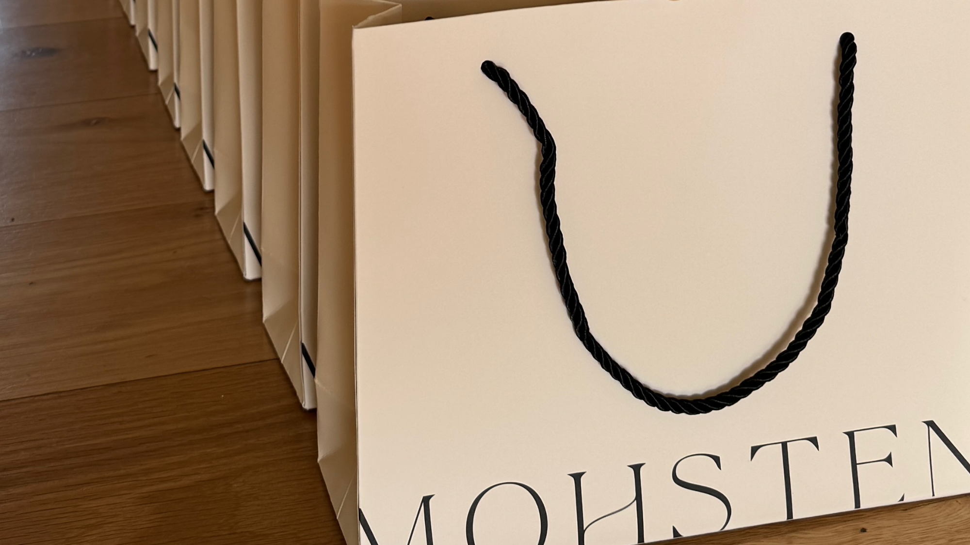 Row of beige gift bags with black handles on a wooden floor, featuring the brand 'MOHSEN'.