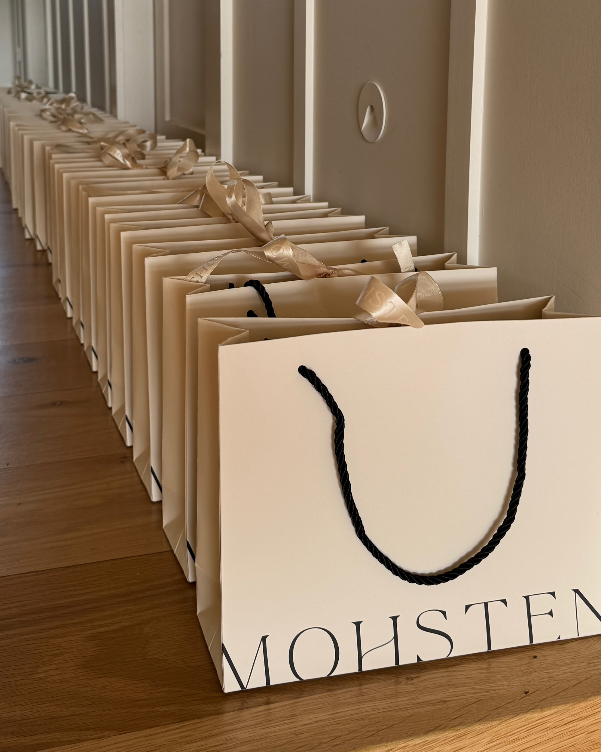 Row of beige gift bags with black handles on a wooden floor, featuring the brand 'MOHSEN'.