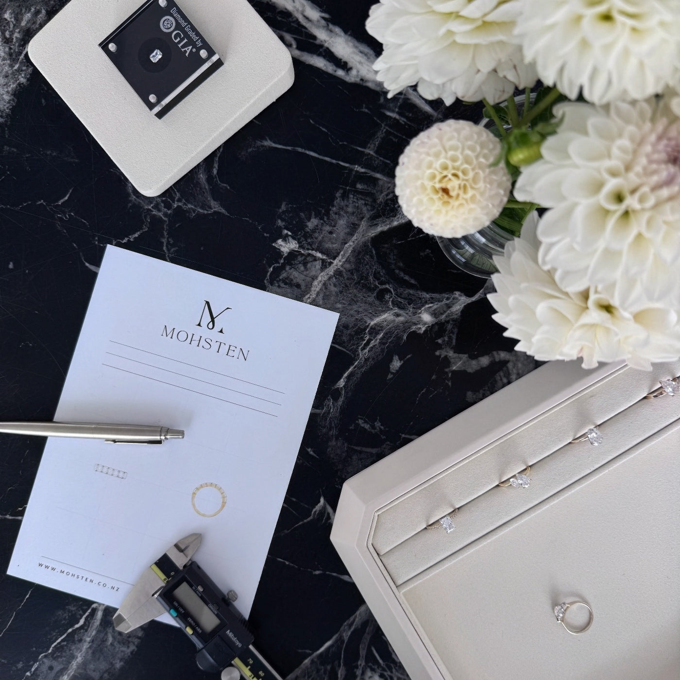 Jewelry box, GIA diamond appraiser, and white flowers on a dark marble surface with 'MOHSTEN' branding.