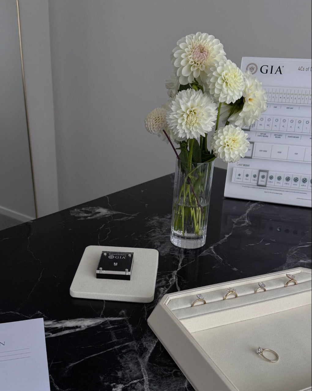 Jewelry display with rings, a vase of flowers, and GIA certificate on a black marble surface.