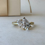 MOHSTEN Cairo pear diamond Toi et Moi engagement ring in 18ct yellow gold in a close-up studio perspective view, highlighting the asymmetrical pairing of the two pear-cut diamonds against a minimalist warm background.