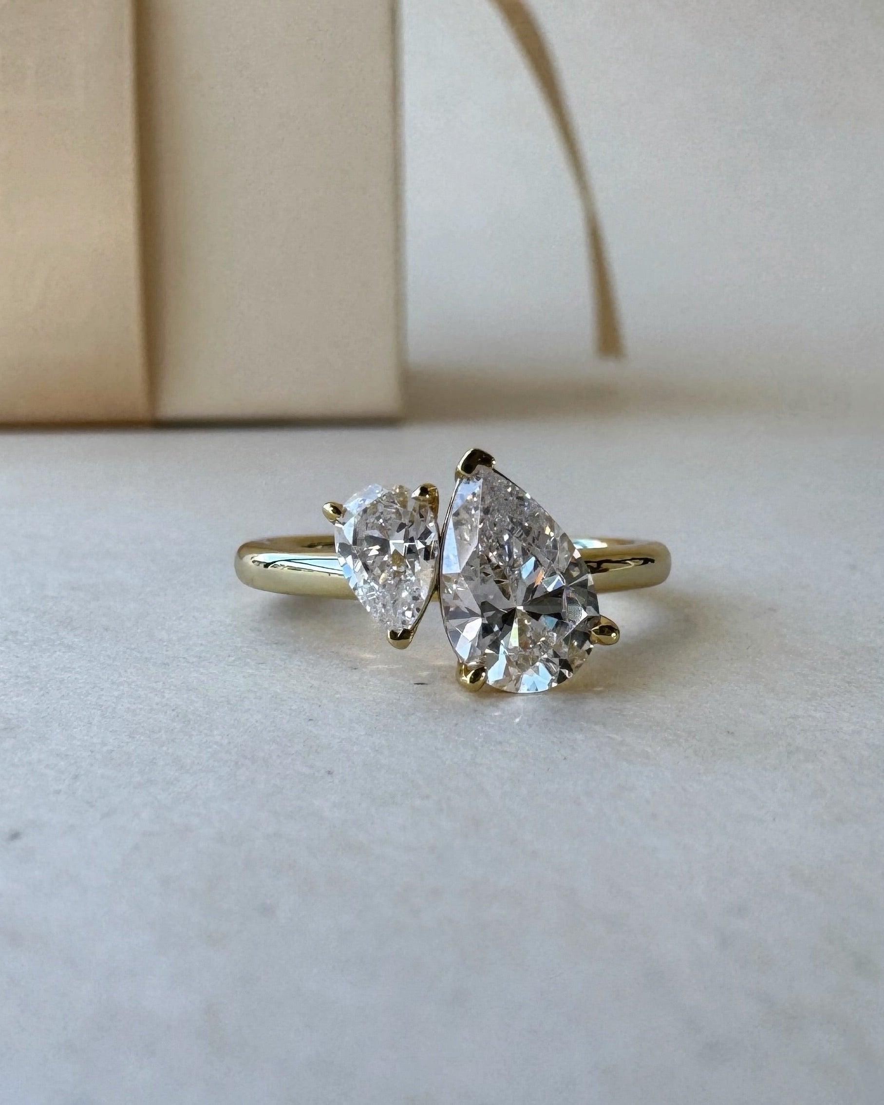 MOHSTEN Cairo pear diamond Toi et Moi engagement ring in 18ct yellow gold in a close-up studio perspective view, highlighting the asymmetrical pairing of the two pear-cut diamonds against a minimalist warm background.