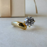 MOHSTEN Granada asymmetric round diamond solitaire engagement ring in 18ct yellow gold, showcasing a three-quarter studio perspective view of the sculptural, wide-swept band and offset round brilliant centre stone resting on a neutral surface.