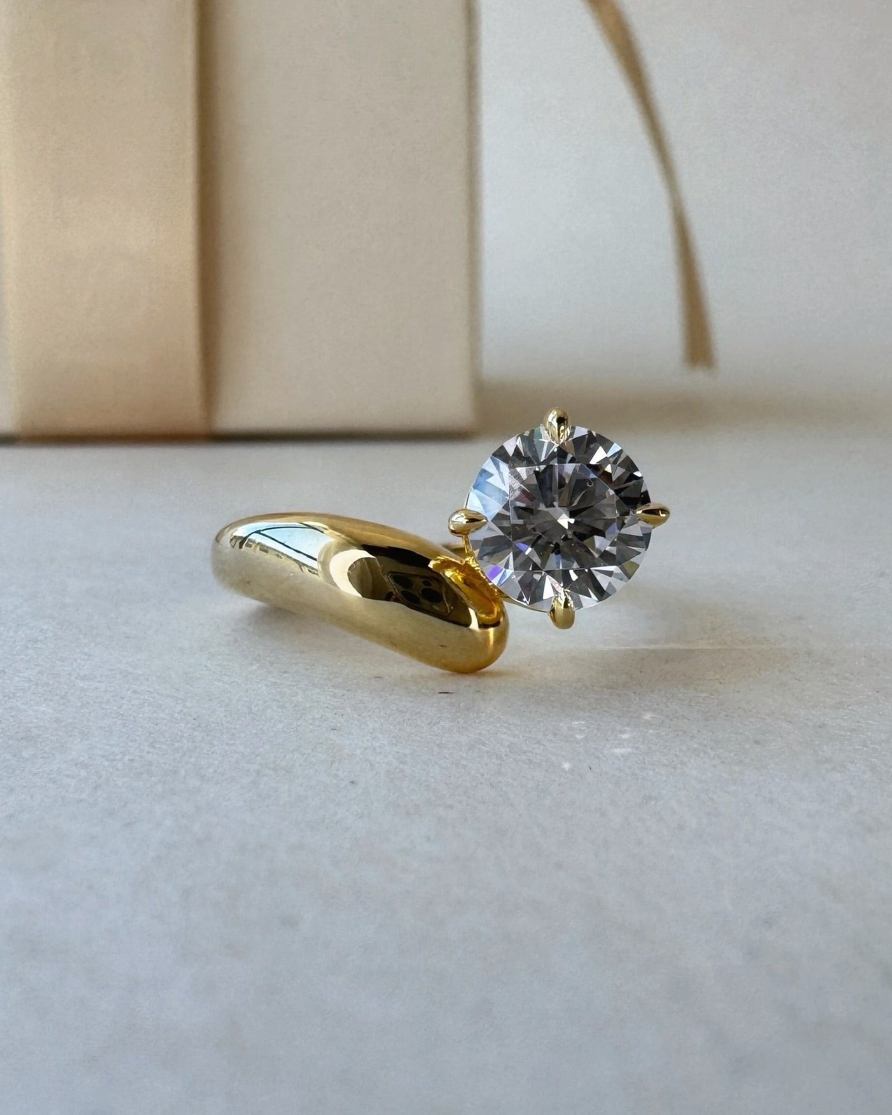 MOHSTEN Granada asymmetric round diamond solitaire engagement ring in 18ct yellow gold, showcasing a three-quarter studio perspective view of the sculptural, wide-swept band and offset round brilliant centre stone resting on a neutral surface.