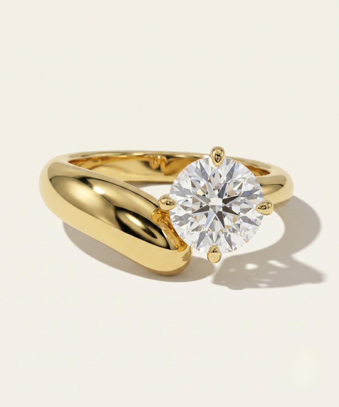 MOHSTEN Granada asymmetric round diamond solitaire engagement ring in 18ct yellow gold showcasing an offset round brilliant diamond on a bold, organic sweeping band casting a soft shadow on a warm studio background.