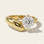 MOHSTEN Granada asymmetric round diamond solitaire engagement ring in 18ct yellow gold showcasing an offset round brilliant diamond on a bold, organic sweeping band casting a soft shadow on a warm studio background.