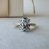 MOHSTEN Osaka oval diamond solitaire engagement ring in 18ct yellow gold showcasing a face-on studio view of the premium oval brilliant centre stone and minimalist 4-prong basket setting resting on a textured surface.
