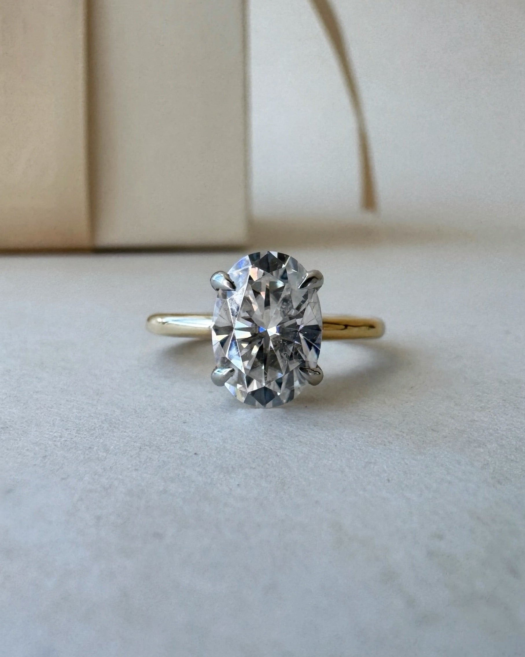 MOHSTEN Osaka oval diamond solitaire engagement ring in 18ct yellow gold showcasing a face-on studio view of the premium oval brilliant centre stone and minimalist 4-prong basket setting resting on a textured surface.
