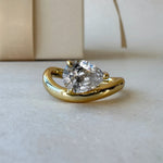 MOHSTEN Venice pear diamond solitaire engagement ring in 18ct yellow gold, showcasing a close-up studio perspective view of the east-west oriented pear diamond and fluid sweeping band next to a luxury gift box.