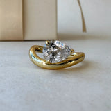 MOHSTEN Venice pear diamond solitaire engagement ring in 18ct yellow gold, showcasing a close-up studio perspective view of the east-west oriented pear diamond and fluid sweeping band next to a luxury gift box.