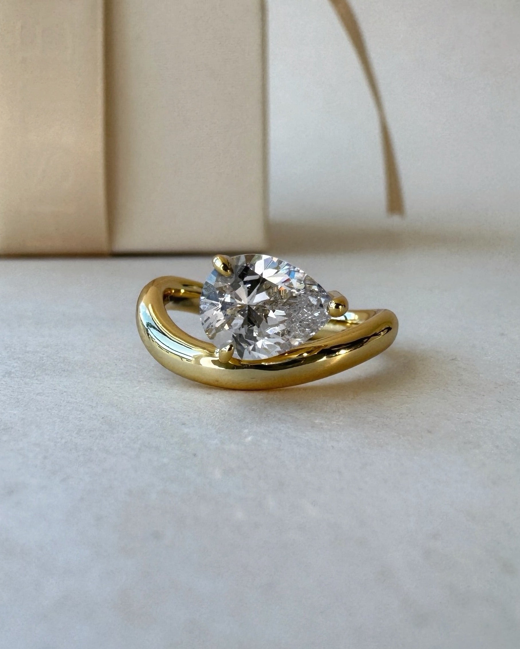 MOHSTEN Venice pear diamond solitaire engagement ring in 18ct yellow gold, showcasing a close-up studio perspective view of the east-west oriented pear diamond and fluid sweeping band next to a luxury gift box.