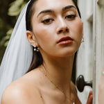 MOHSTEN bride wearing a white bridal veil and strapless white wedding dress, leaning against a weathered wooden door with a black doorknob. She is wearing a delicate gold necklace with a keshi pearl pendant