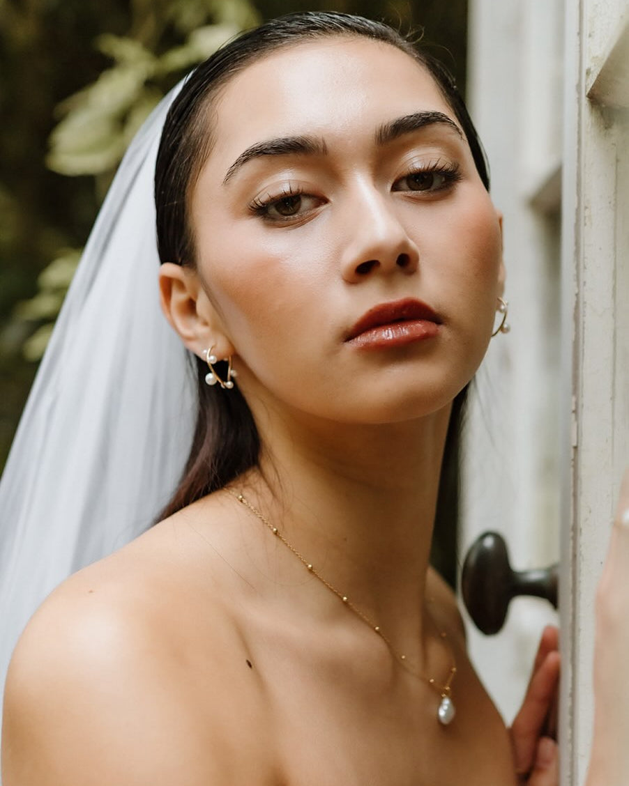 MOHSTEN bride wearing a white bridal veil and strapless white wedding dress, leaning against a weathered wooden door with a black doorknob. She is wearing a delicate gold necklace with a keshi pearl pendant