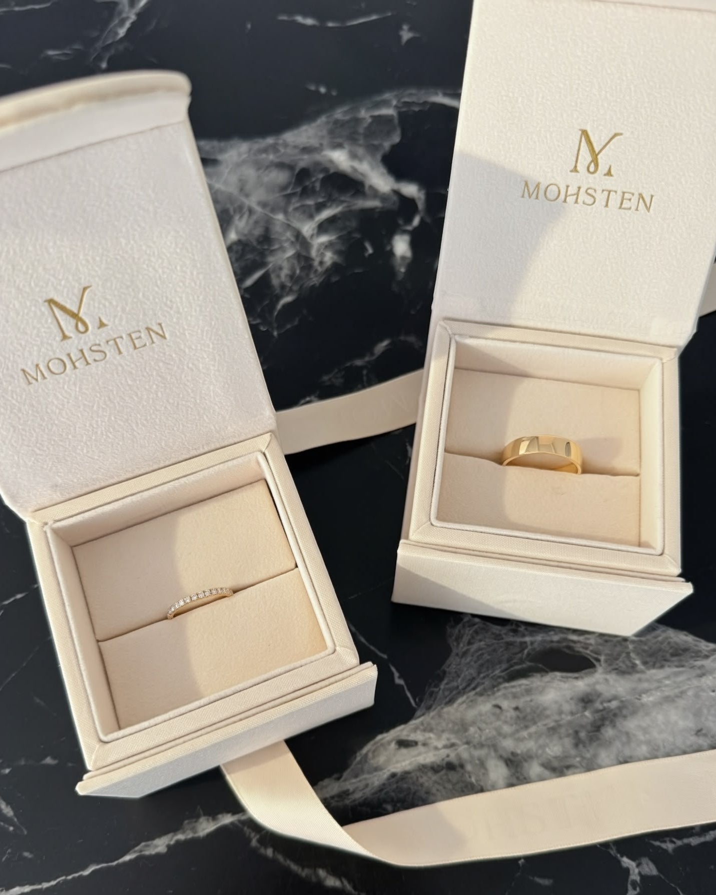 Two open jewelry boxes with rings inside on a black marble surface, branded 'MOHSTEN'.